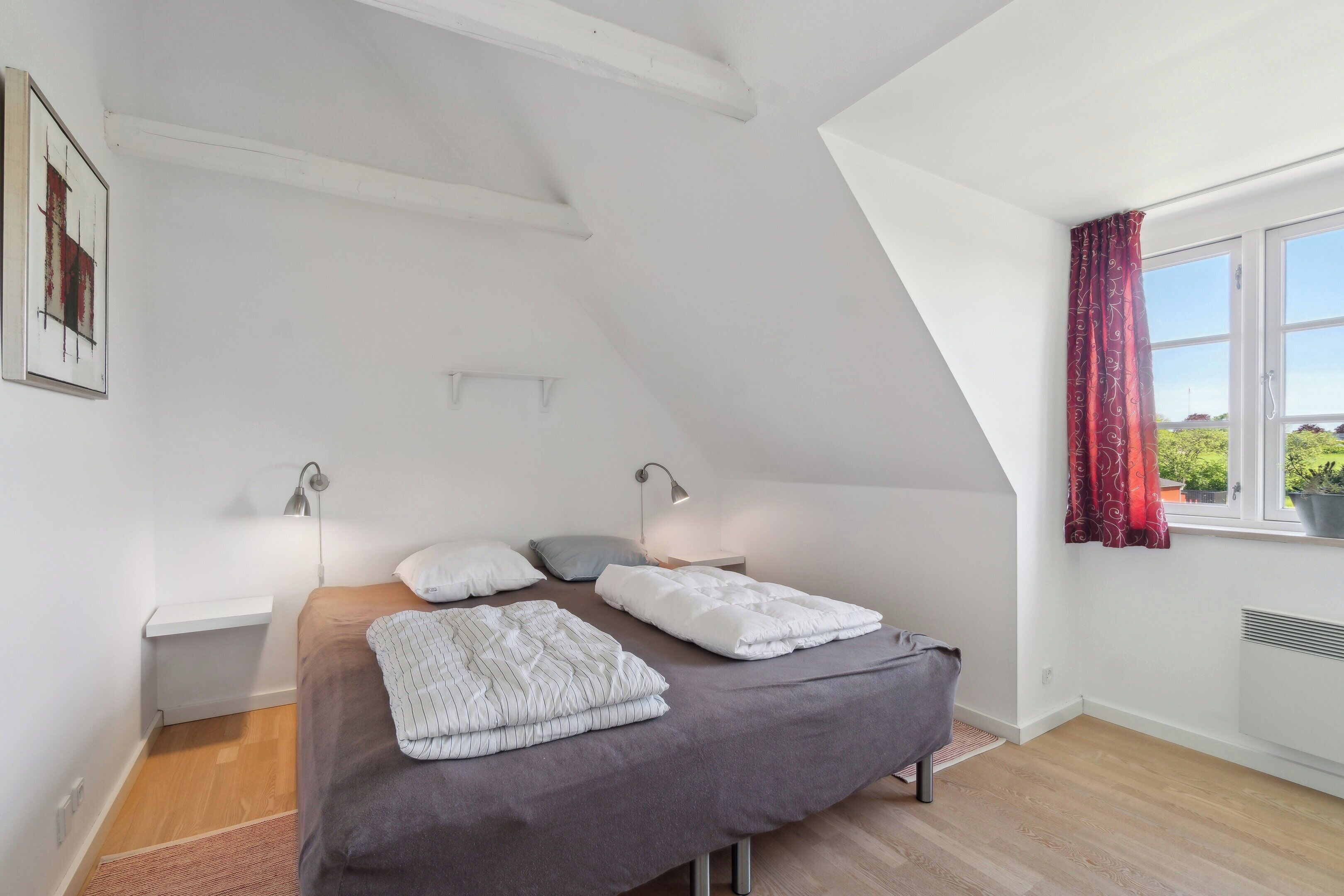 Une chambre avec un plafond en pente, un lit avec deux oreillers, une table de chevet avec une lampe et une fenêtre avec des rideaux rouges.