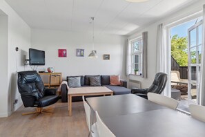 A modern living room with a sofa, chairs, a dining table, a TV, and large windows.
