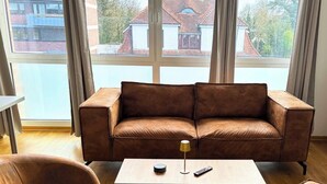 A brown leather sofa with a remote control and a lamp on a wooden table in a room with a large window and a radiator. - Studio Apartment 'Panorama' with Wi-Fi (Lingen (Ems))
