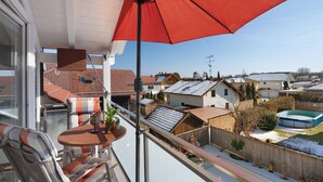 Ein Balkon mit rotem Sonnenschirm, einem Tisch mit Stühlen und Blick auf Nachbarhäuser sowie ein Schwimmbad.
