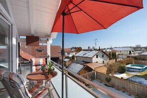 Un balcón con una sombrilla roja, una mesa con sillas y vistas a casas vecinas y una piscina.