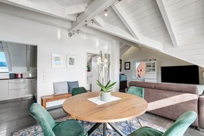 Ein modernes Wohnzimmer mit einem runden Holztisch, grünen Polsterstühlen, einem braunen Sofa und einem Fernseher.