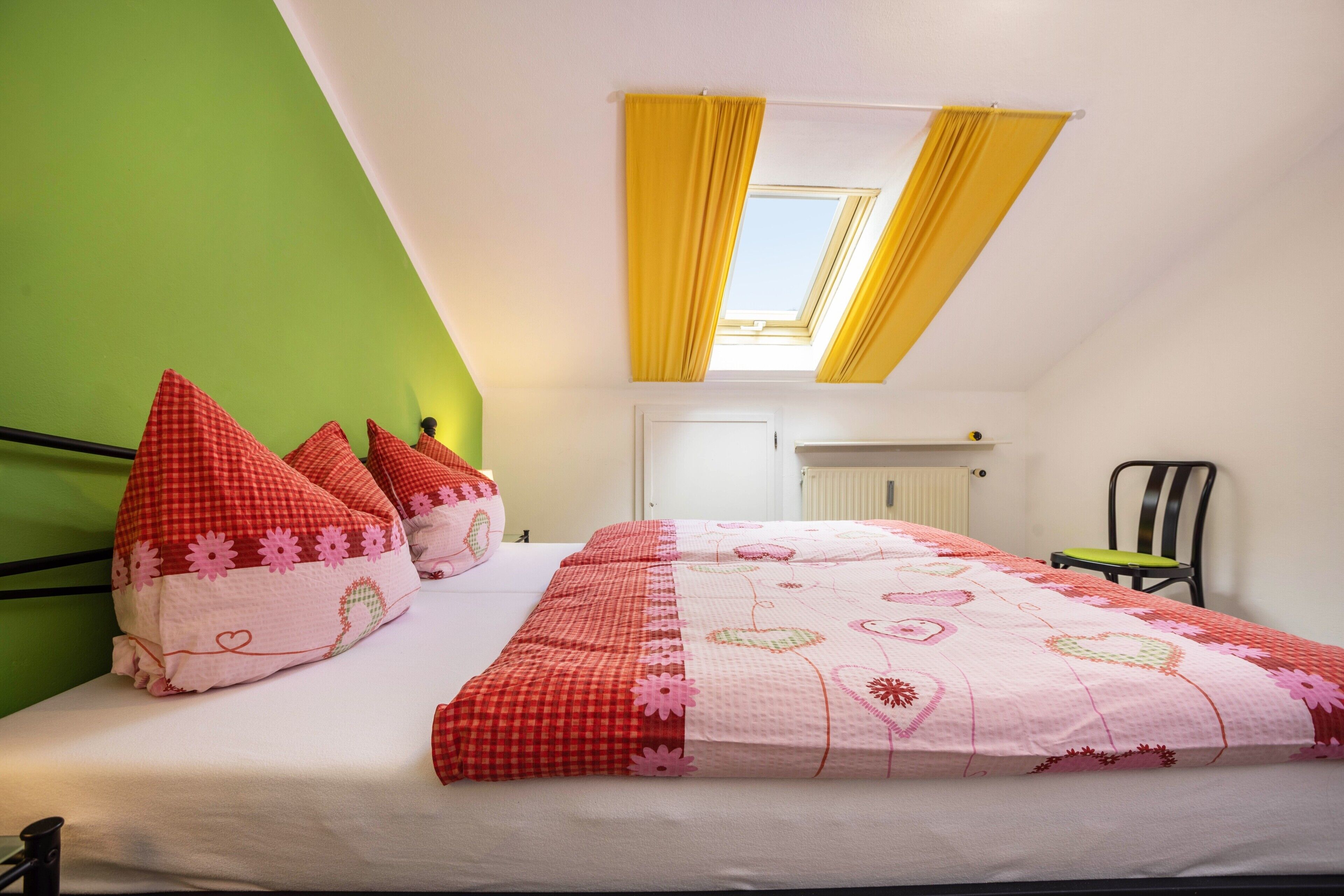 Un dormitorio con cama, paredes verdes, tragaluz y una silla.