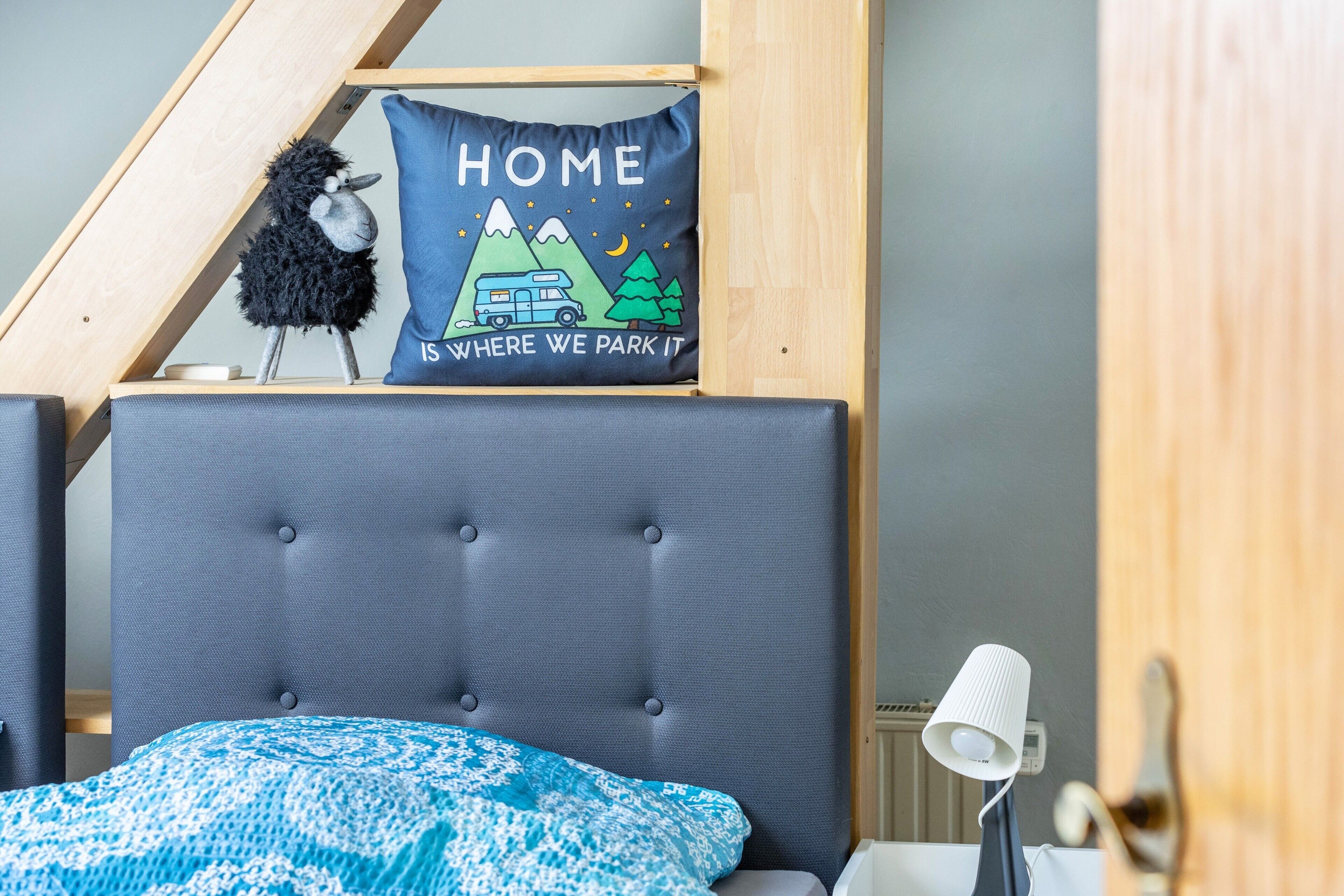 Een slaapkamer met een grijs gestoffeerd hoofdeinde, een blauw bed met een patroon, een kussen met een reisontwerp en een klein zwart pluche beestje.