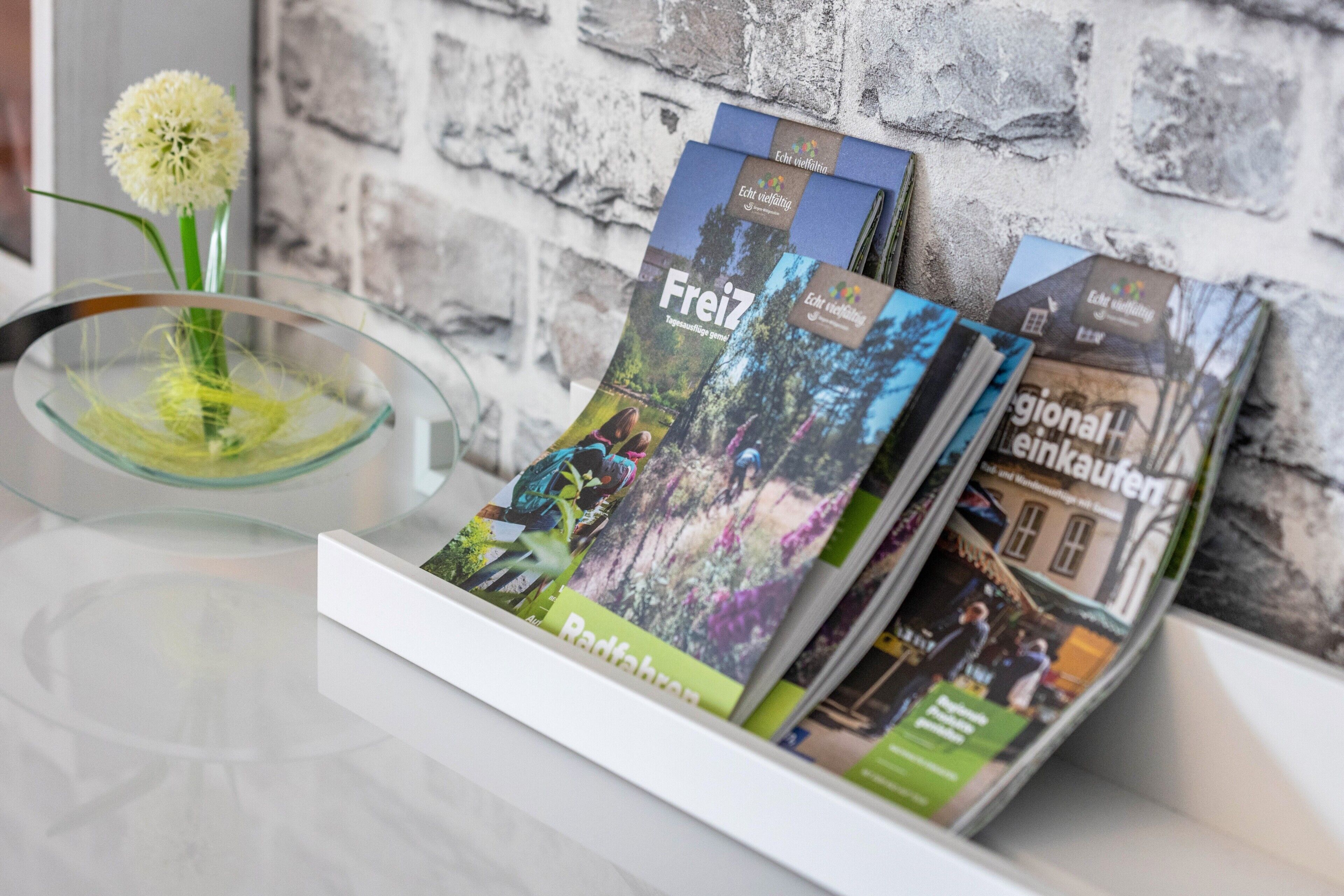 Ein Zeitschriftenständer mit Reisemagazinen, eine Schale mit einer Blume und eine Backsteinwand im Hintergrund.