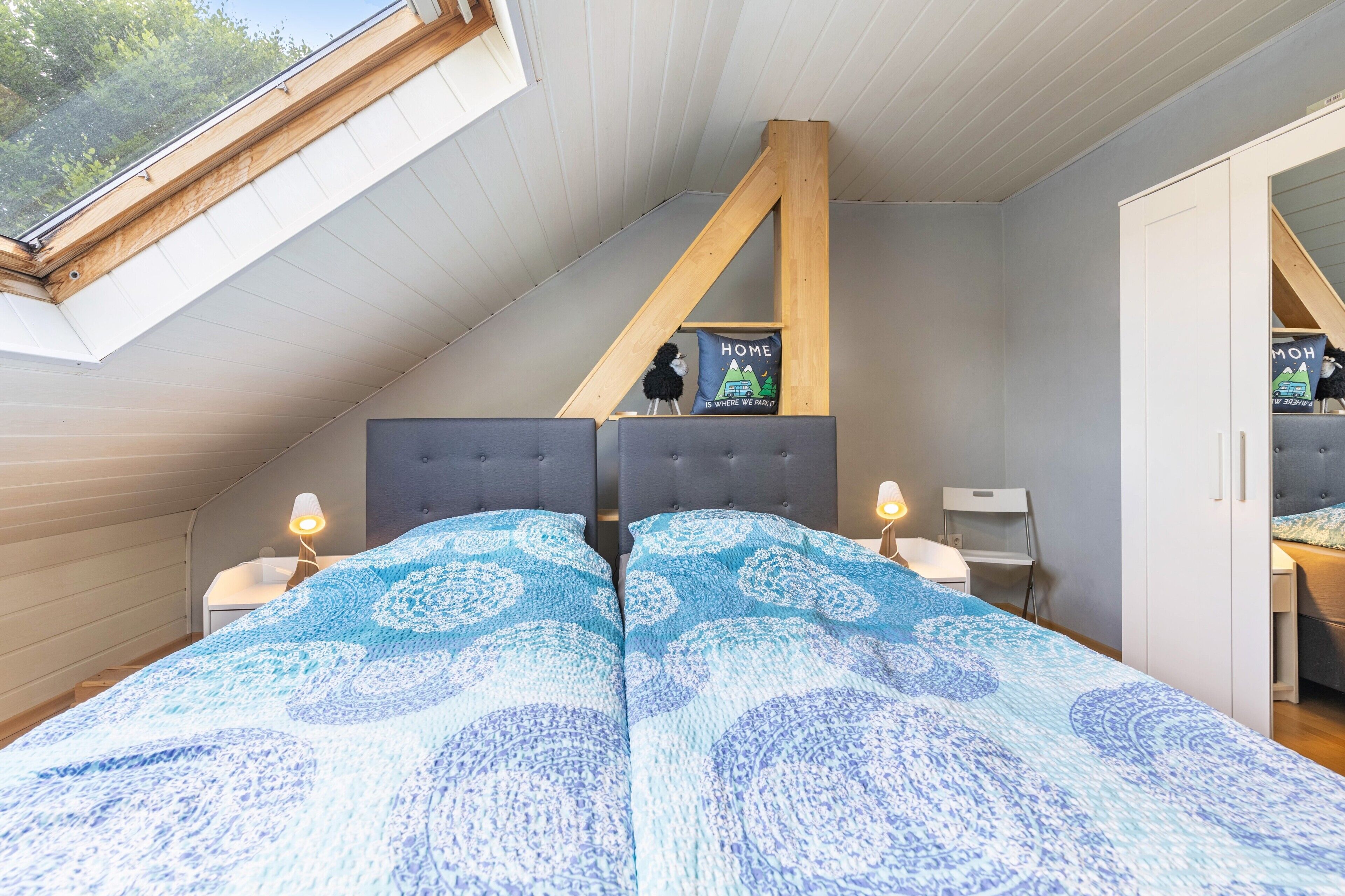 Une chambre avec un plafond en pente, un lit recouvert d’une couvre-lit à motifs, un petit bureau et une étagère fixée au mur, agrémentée d’un coussin.