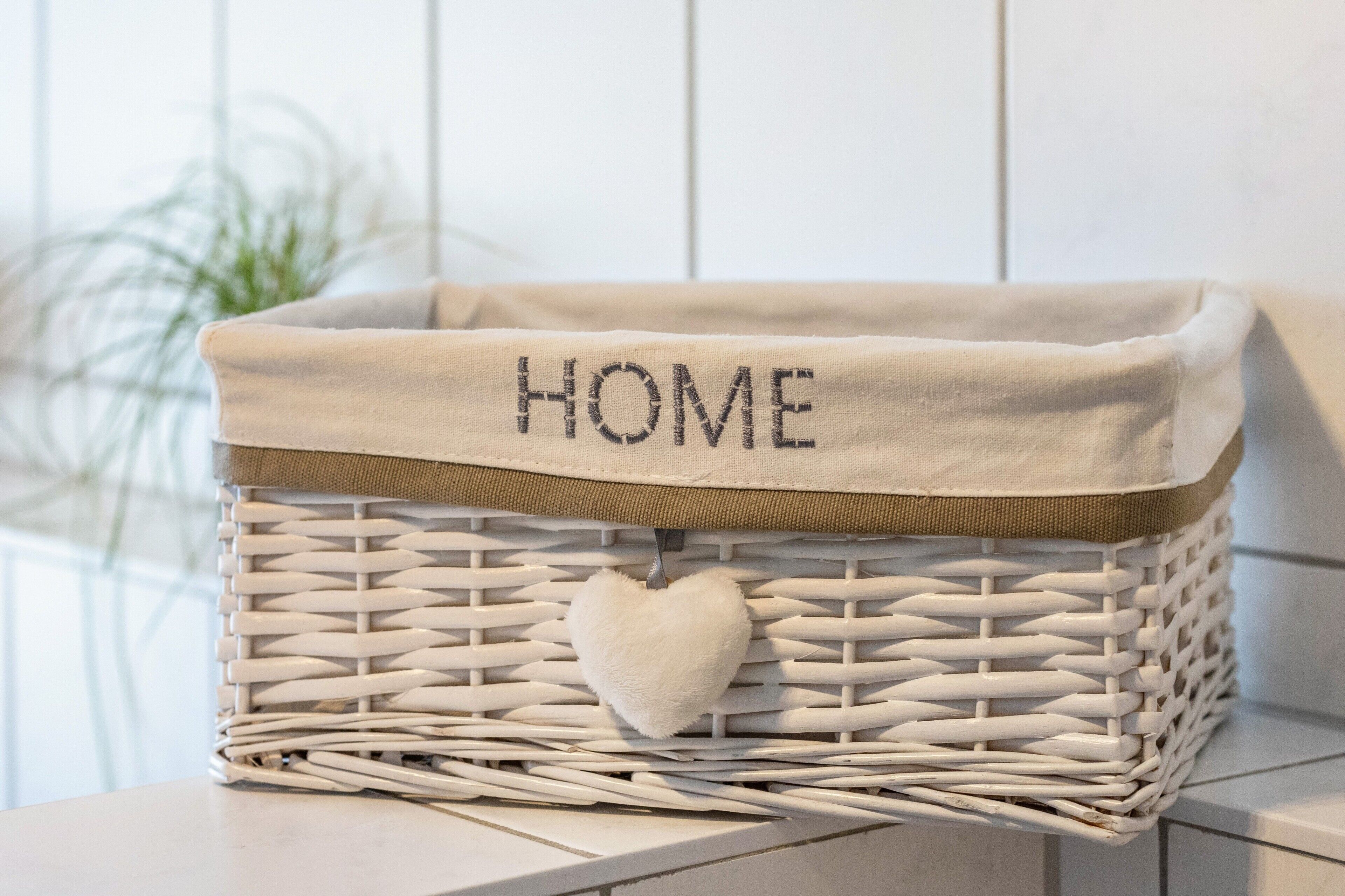 A white wicker basket with a beige fabric top labeled 'HOME' and a white heart-shaped object inside.