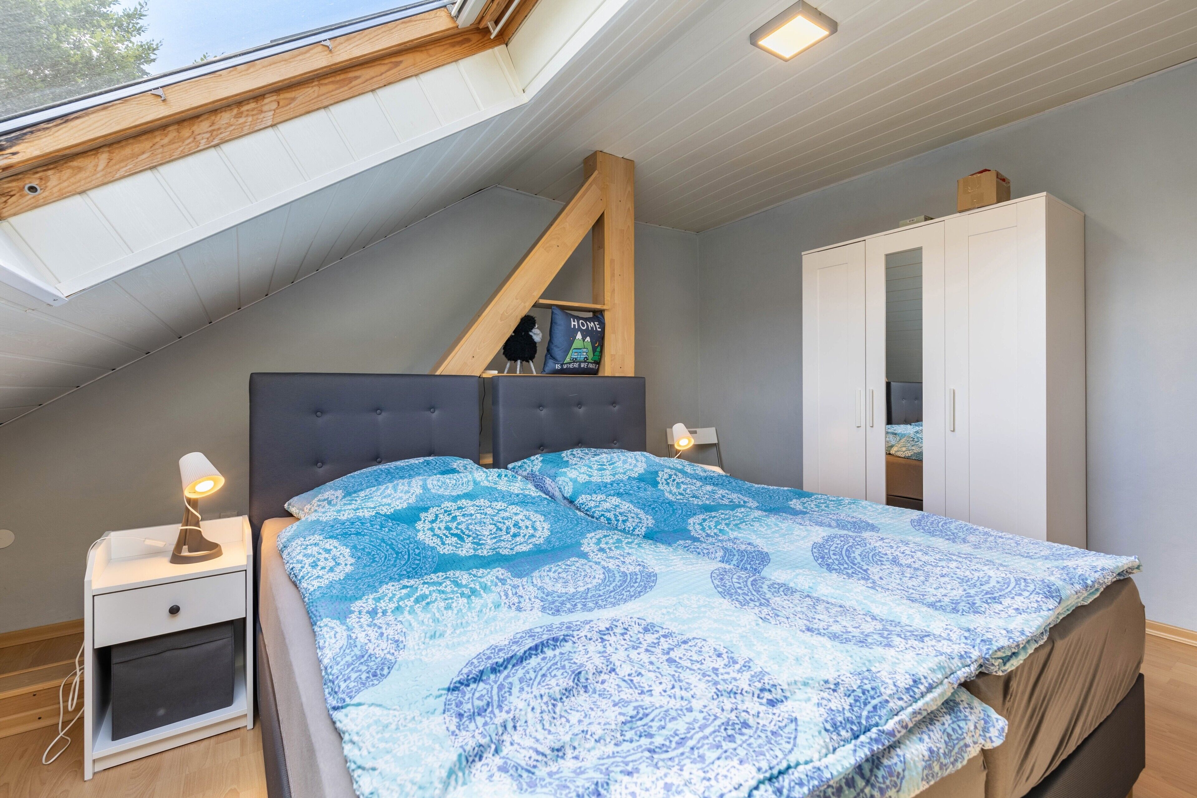 A bedroom with a sloped ceiling, a bed with a patterned bedspread, a wardrobe, and a nightstand with a lamp.