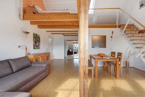 A spacious living area with a wooden staircase, a sofa, a dining table with chairs, and a large window.