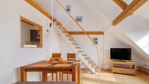 Un espai tipus loft amb una escala de fusta, una taula de dinar, una banca, una televisió i un gran finestral.