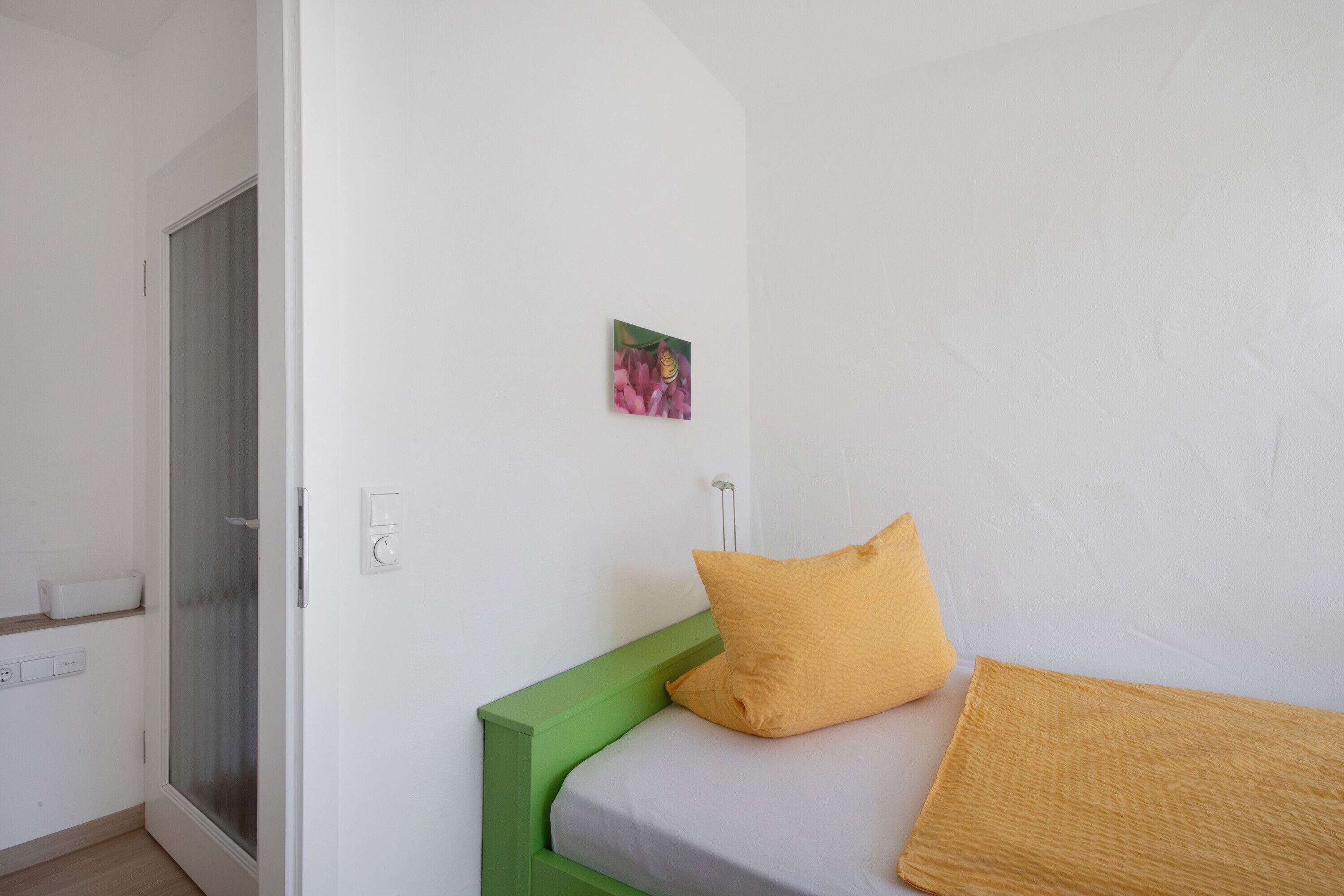 Une chambre à coucher avec un lit, une tête de lit verte, un seul oreiller jaune, un petit tableau accroché au mur et une porte dotée d’un panneau en verre dépoli.