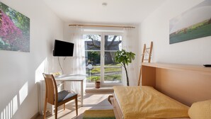 Ein Schlafzimmer mit einem Bett, einem Schreibtisch, einem Stuhl, einem Fernseher und einem Fenster mit Blick auf Grünflächen.