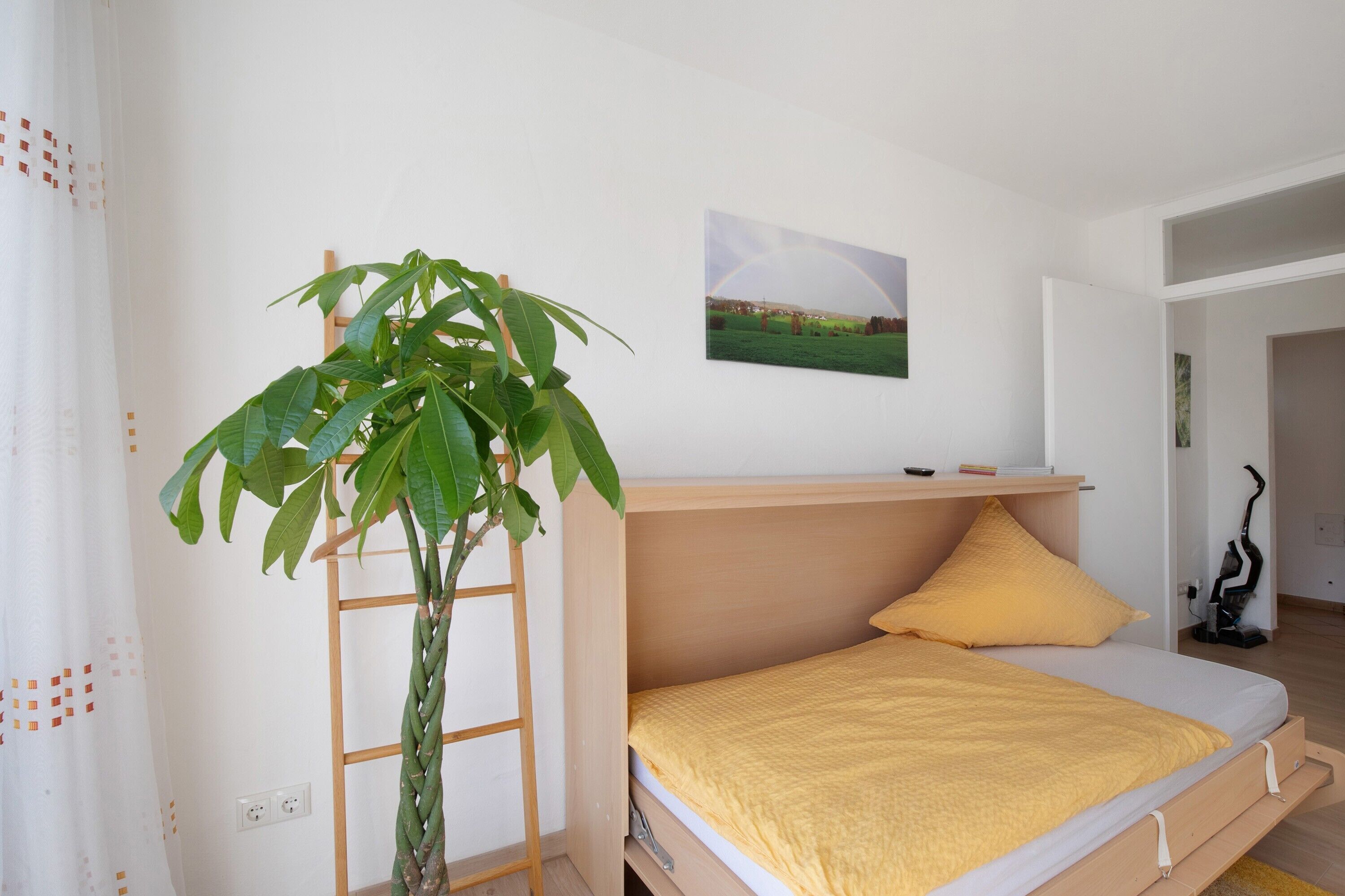 A bedroom with a bed, a plant, a ladder, a painting, and a vacuum cleaner.