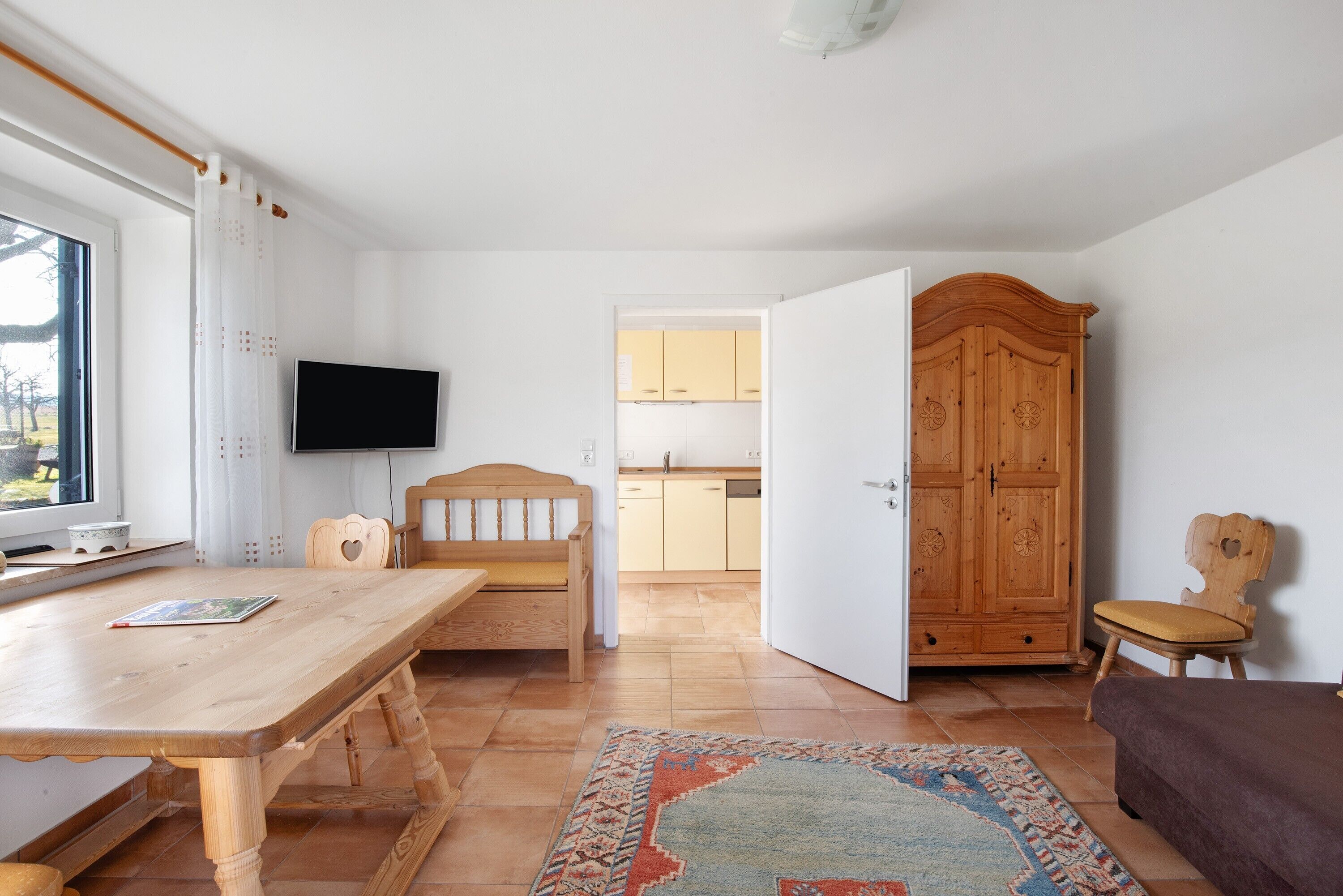 Una stanza con un tavolo di legno, una panca, una televisione, un armadio e vista sulla cucina.