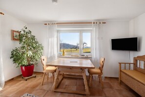Ein Essbereich mit Holztisch, Stühlen, einer Zimmerpflanze, einem Fernseher und Blick ins Freie.