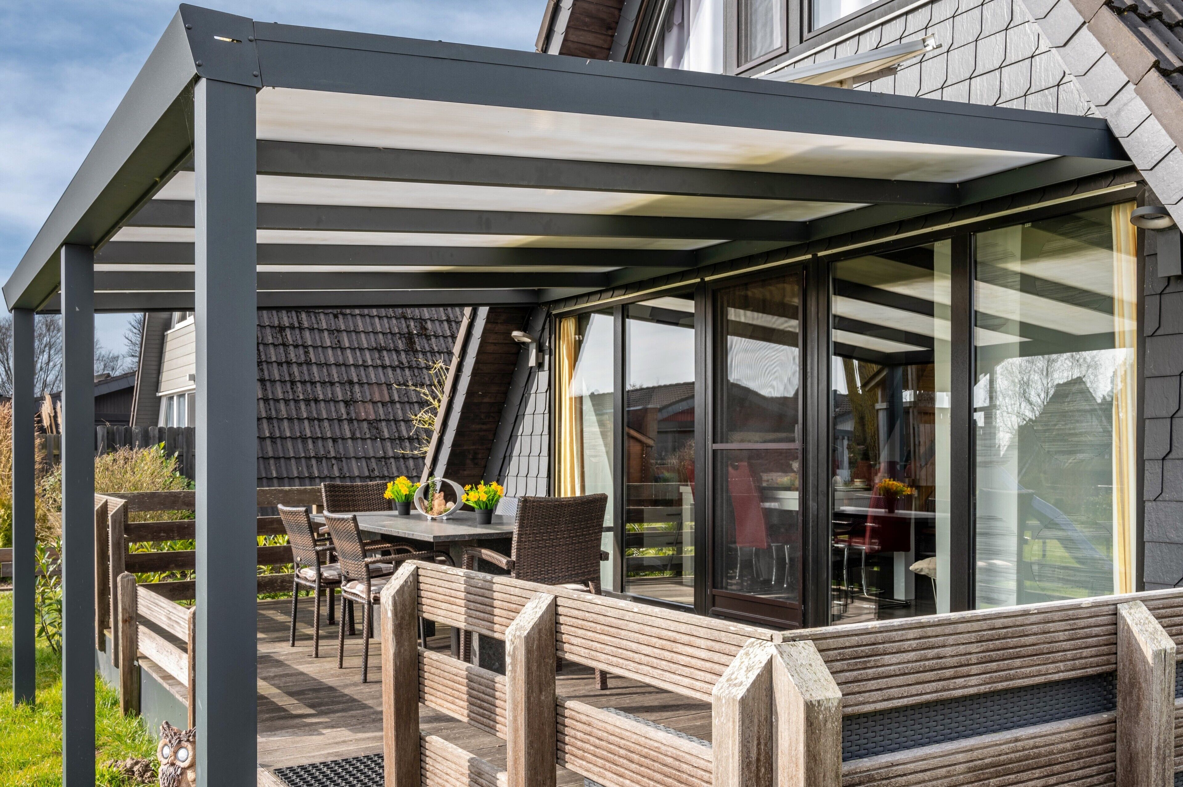 A covered outdoor seating area with a table and chairs, a bench, and a view of the house interior through glass doors.