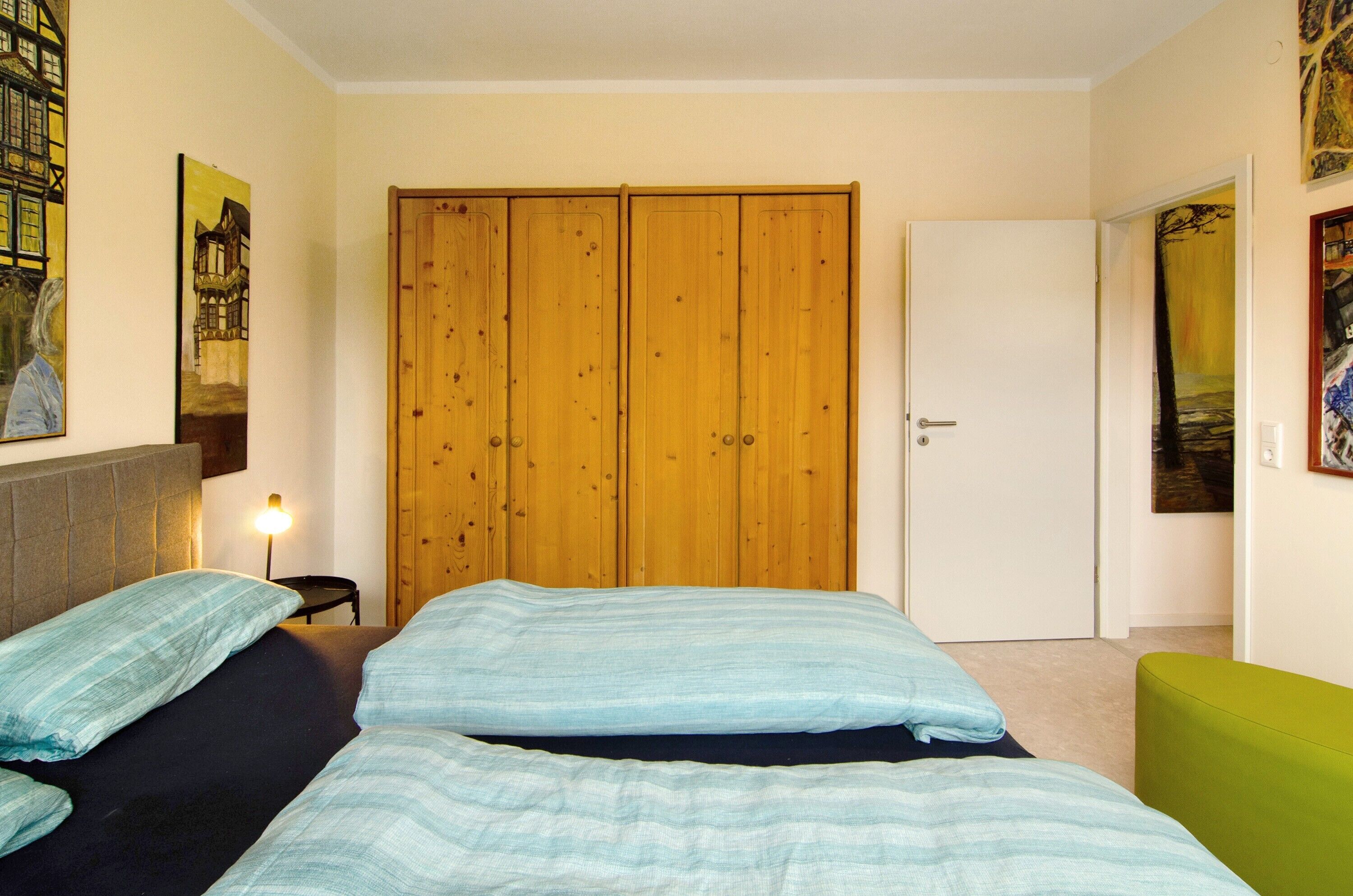 Una camera da letto con due letti, un'armadio in legno, una sedia verde e opere d'arte alle pareti.