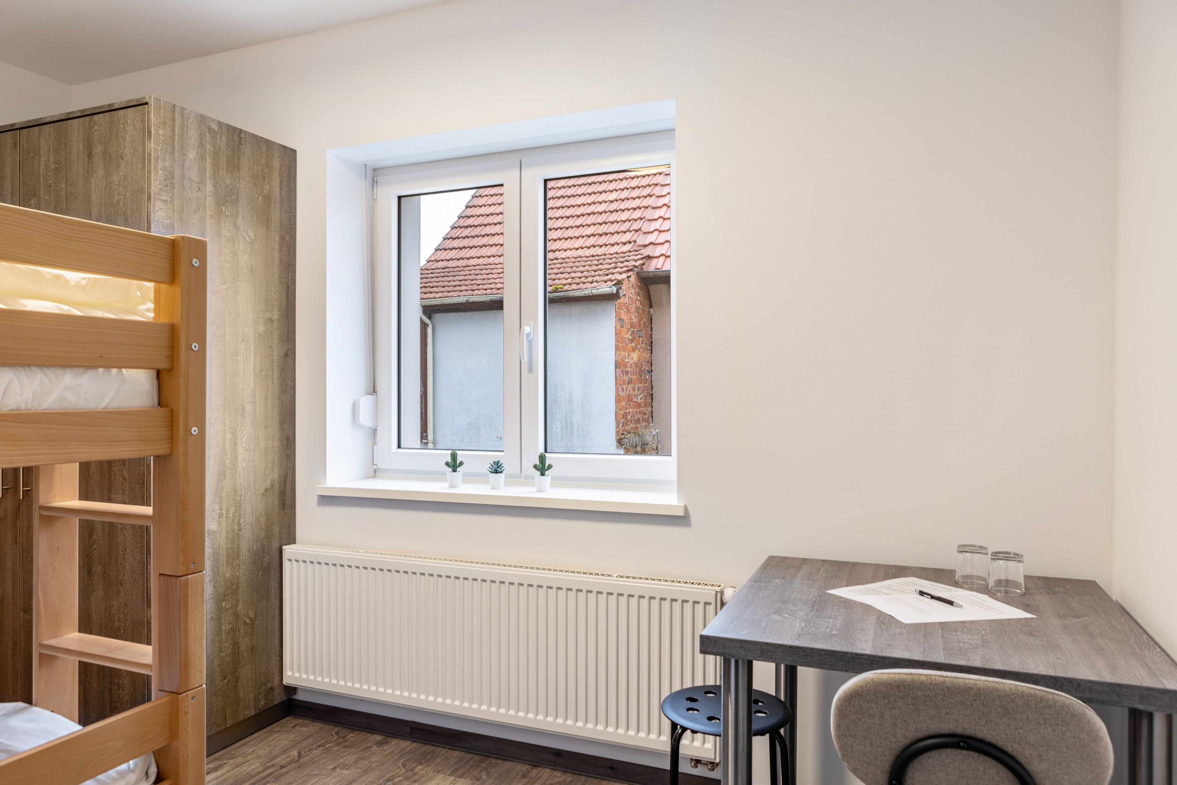 Ein Zimmer mit einem Etagenbett, einem Tisch mit Stuhl, einem Fenster mit Blick auf ein Gebäude und einem Heizkörper.