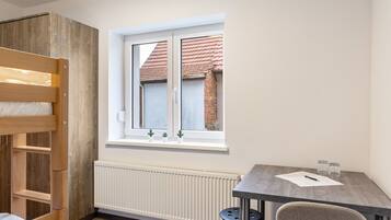 Ein Zimmer mit einem Etagenbett, einem Tisch mit Stuhl, einem Fenster mit Blick auf ein Gebäude und einem Heizkörper.