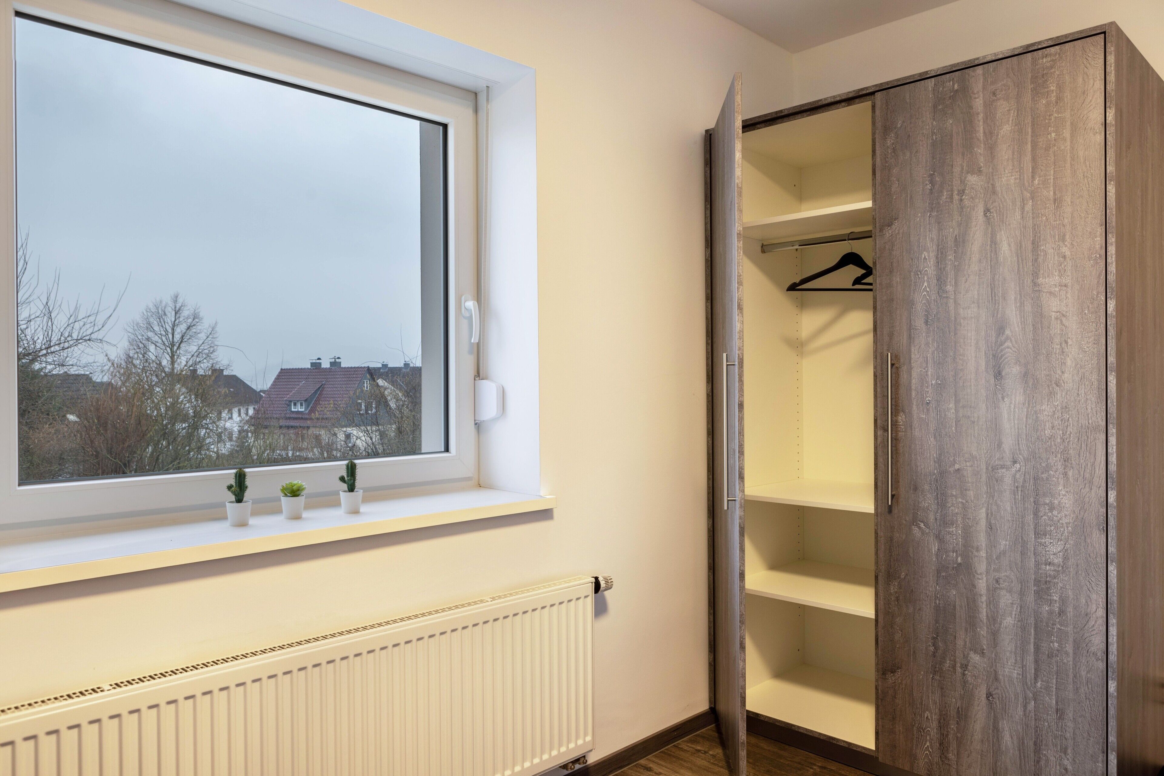 Ein Zimmer mit Fenster, Heizkörper, einem Holzschrank und einem kleinen Regal mit Pflanzen.