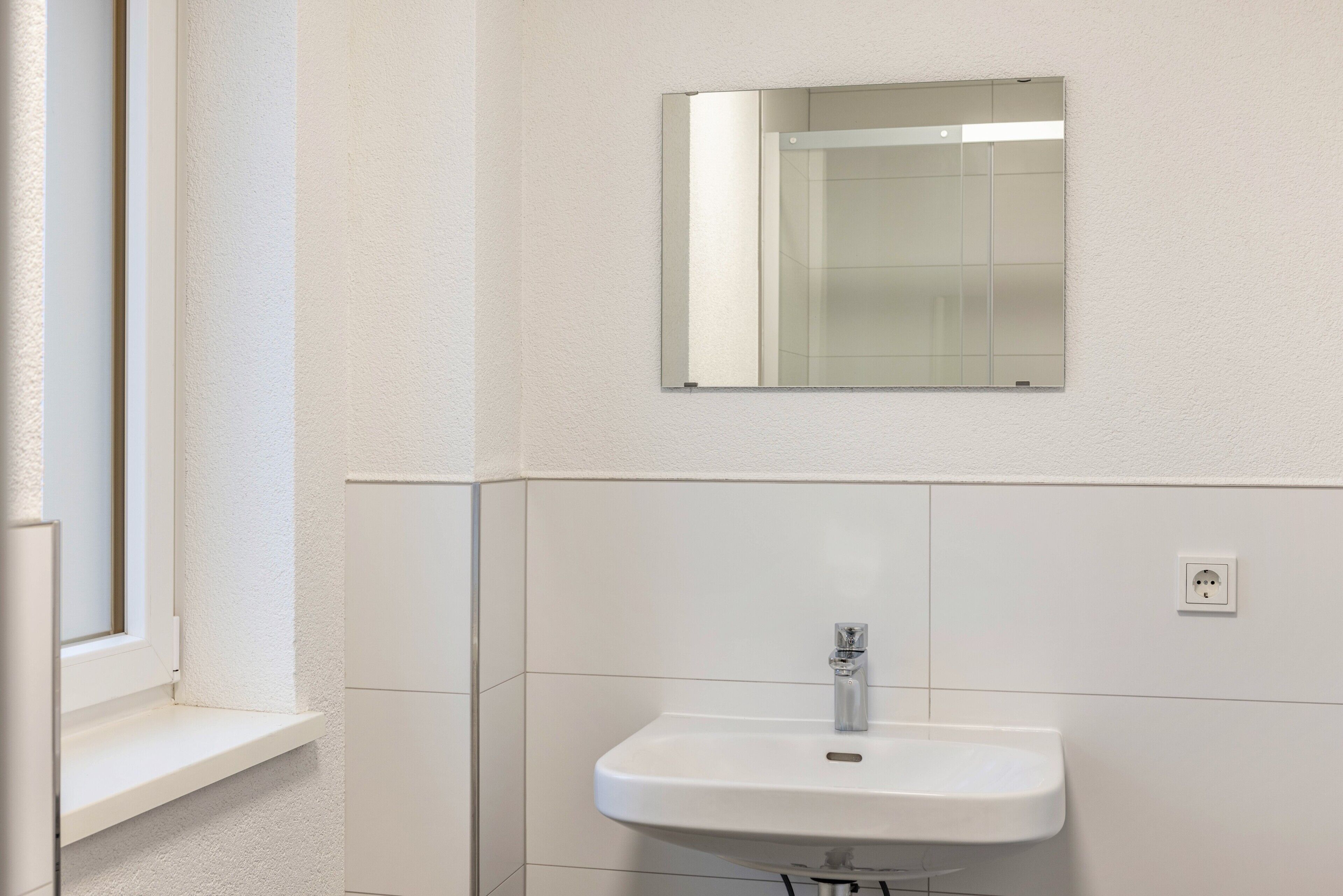 Un bagno con un lavandino bianco, uno specchio sopra e una finestra a sinistra.