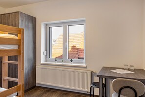 Une pièce avec un lit superposé, une fenêtre donnant sur des immeubles, un radiateur, une petite table avec des papiers et une chaise.