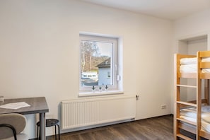 Una habitación con ventana, radiador, literas, escritorio y silla.