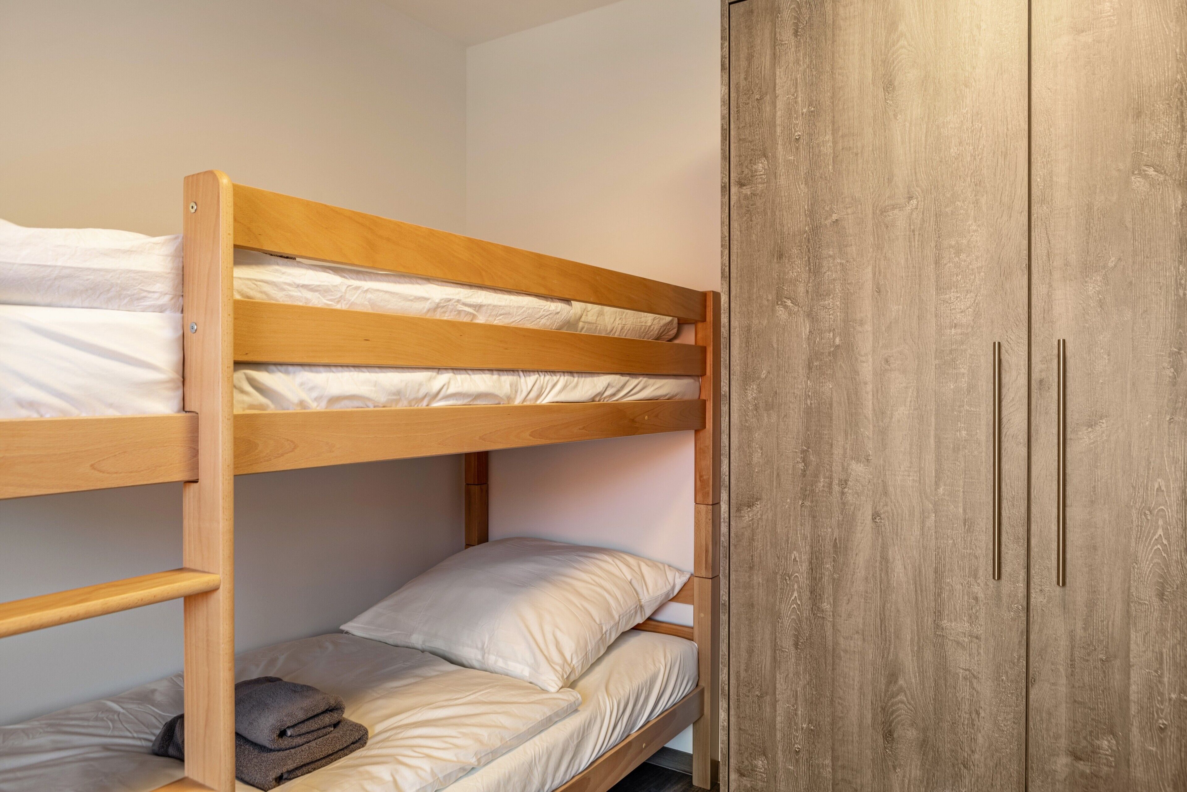 A bunk bed with wooden headboard and drawers, a wardrobe with two doors, and a grey towel on the lower bunk.