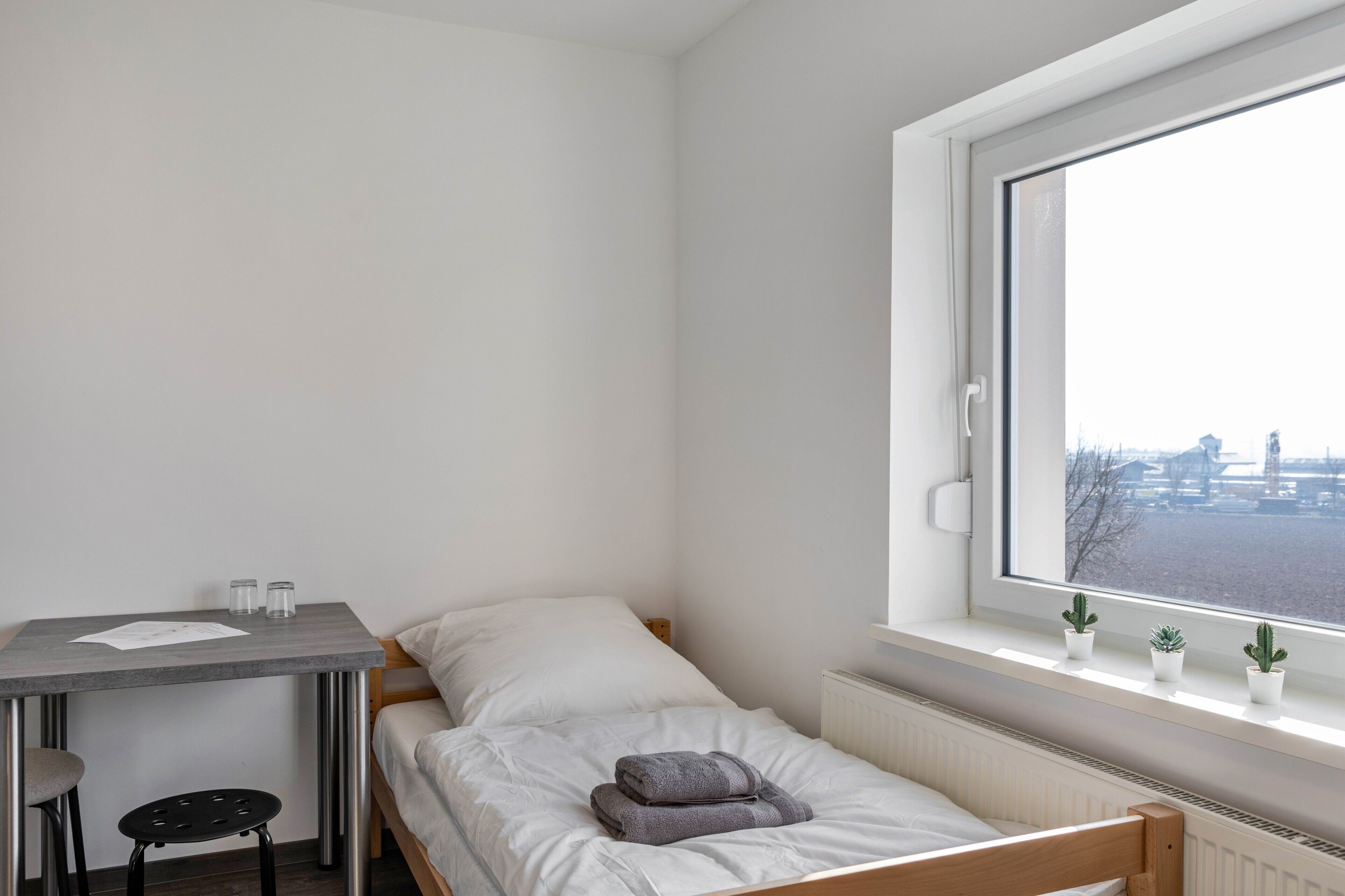 Une petite pièce propre, avec un lit simple, une table et une chaise. Il y a une fenêtre donnant sur des immeubles et un radiateur.