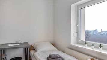 Une petite pièce propre, avec un lit simple, une table et une chaise. Il y a une fenêtre donnant sur des immeubles et un radiateur.