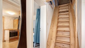 Una escalera de madera que lleva a una habitación con espacio de cocina, una cortina azul y una pared de ladrillo blanco.