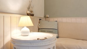 Una mesa redonda blanca con una lámpara, un cojín a rayas con el texto 'deep ocean' y una decoración de barco en la pared.