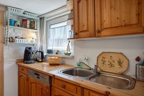 Una cocina con armarios de madera, pared de ladrillo blanco, ventana con cortinas, un fregadero y una cafetera.