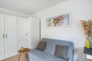 Quarto com sofá cinzento, uma pequena mesa de cabeceira de madeira, guarda-roupa branco e um quadro abstrato emoldurado na parede.
