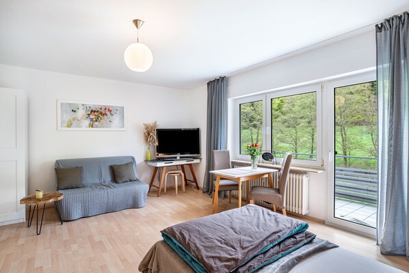 Une chambre moderne équipée d’un lit, d’un canapé, d’une télévision, d’une table à manger et d’une fenêtre donnant sur un paysage verdoyant.
