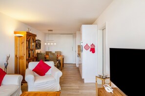 Een woonkamer met een witte bank, rode kussens, een houten kast, een eethoek, en een televisie aan de rechterkant.