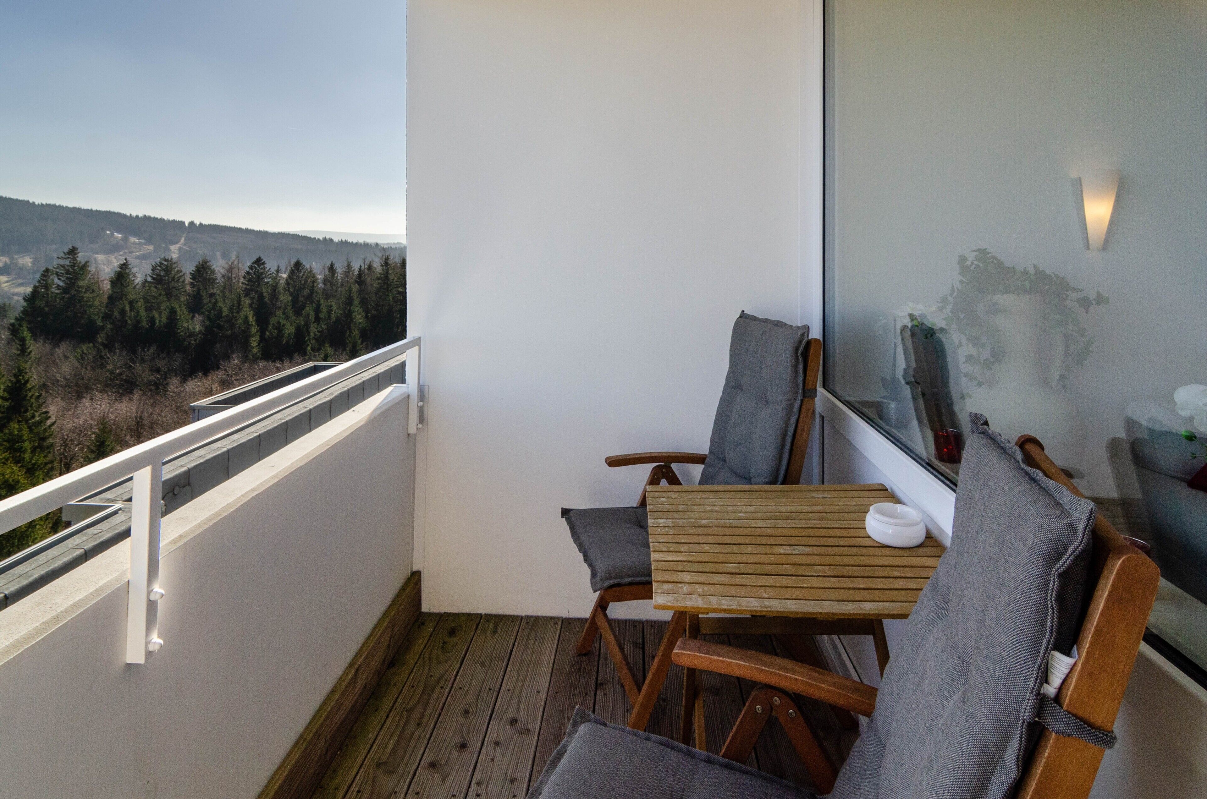 Un balcon donnant sur un paysage boisé, une table en bois, deux chaises et un bol blanc.