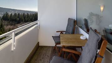 Un balcon donnant sur un paysage boisé, une table en bois, deux chaises et un bol blanc.