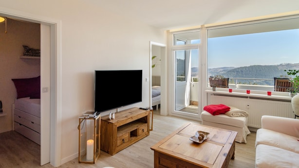 A living room with a TV, wooden furniture, a sofa, and a view of a mountainous landscape through large windows. - Apartment 'Wolke8' with Private Sauna, Stunning Mountain Views, Balcony, Wi-Fi, Parking (Goslar-Hahnenklee)