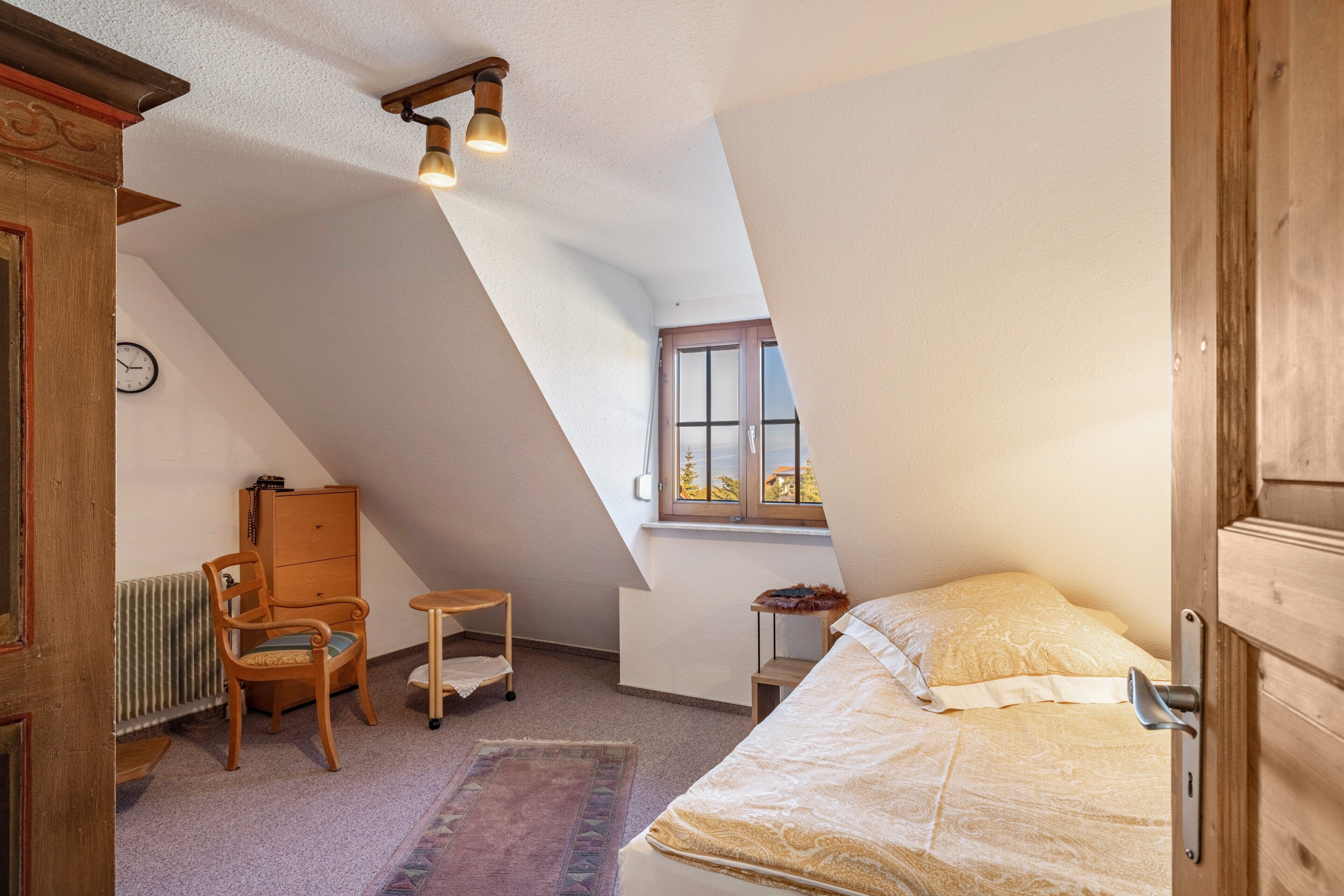 A bedroom with a sloped ceiling, a bed, a chair, a small table, a radiator, and a window.