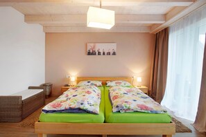 Een tweepersoonsbed met bloemenbeddengoed, een houten hoofdbord, een klein nachtkastje, een aan de muur hangend schilderij en een raam met gordijnen.