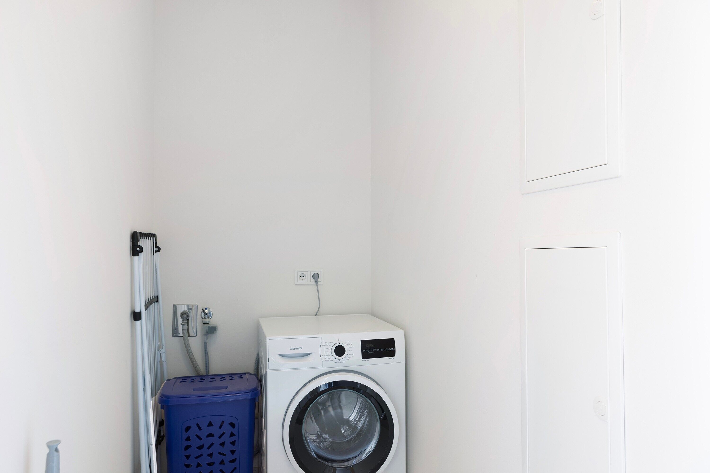 A compact laundry room with a front-loading washing machine, a blue laundry basket, and a metal drying rack.
