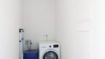 A compact laundry room with a front-loading washing machine, a blue laundry basket, and a metal drying rack.