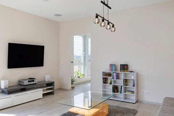 Un salon moderne avec un téléviseur à écran plat, une table basse en verre, une bibliothèque blanche et un luminaire suspendu.