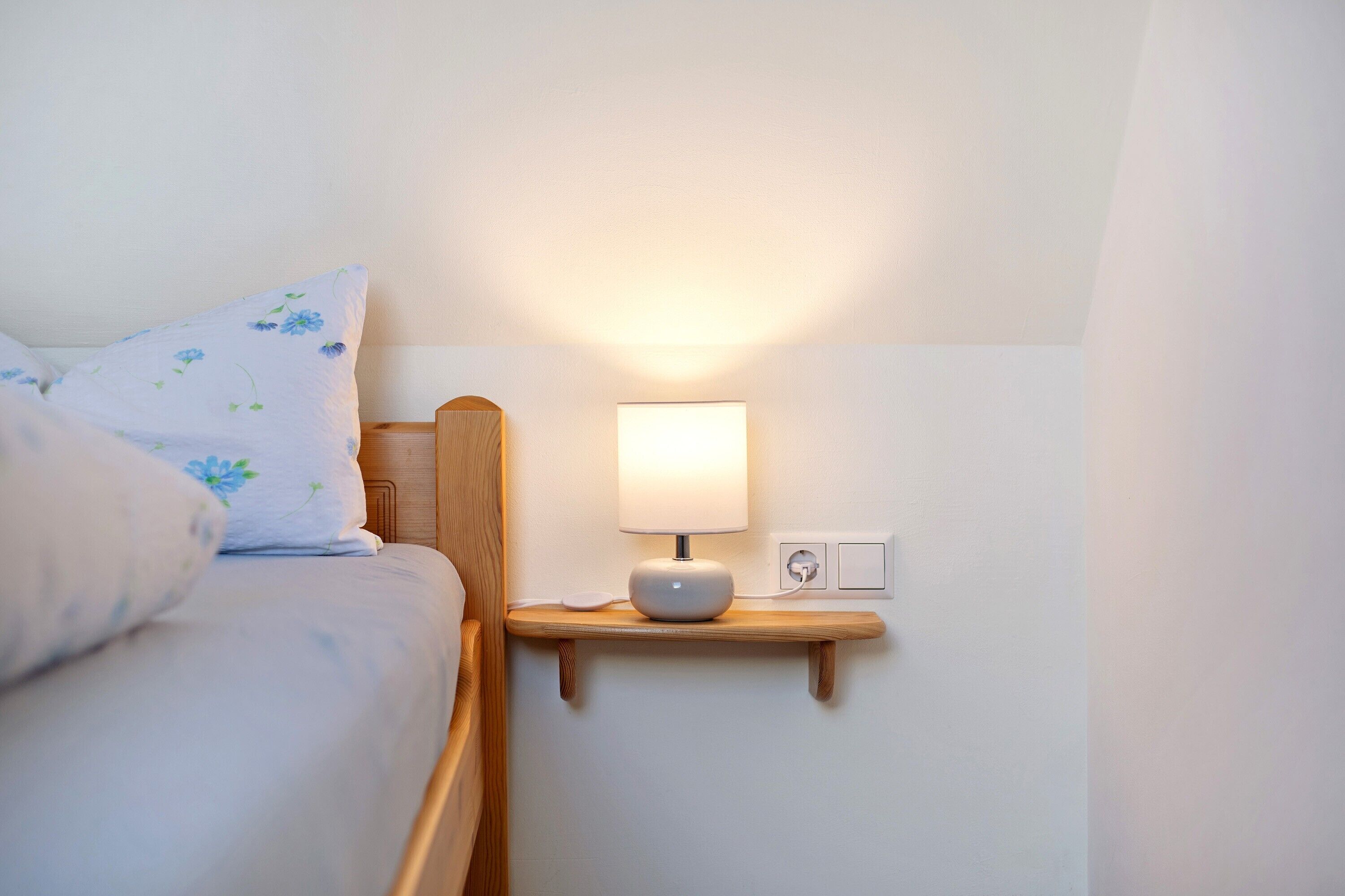 Ein ordentlich bezogenes Bett mit floralen Kissen, ein Nachttisch mit Lampe und eine Wandsteckdose.