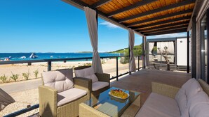 A covered outdoor seating area with wicker furniture and a glass table, overlooking a beach and the sea. - Camp Adriatik (Primosten)