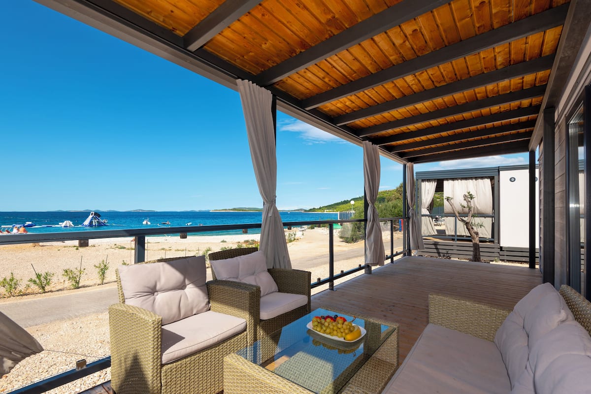 A covered outdoor seating area with wicker furniture and a glass table, overlooking a beach and the sea.
