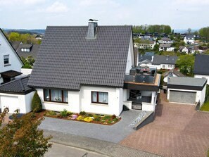 Une maison moderne à deux étages, dotée d’un toit en pente, d’un petit jardin agrémenté de fleurs colorées et d’une allée pavée.