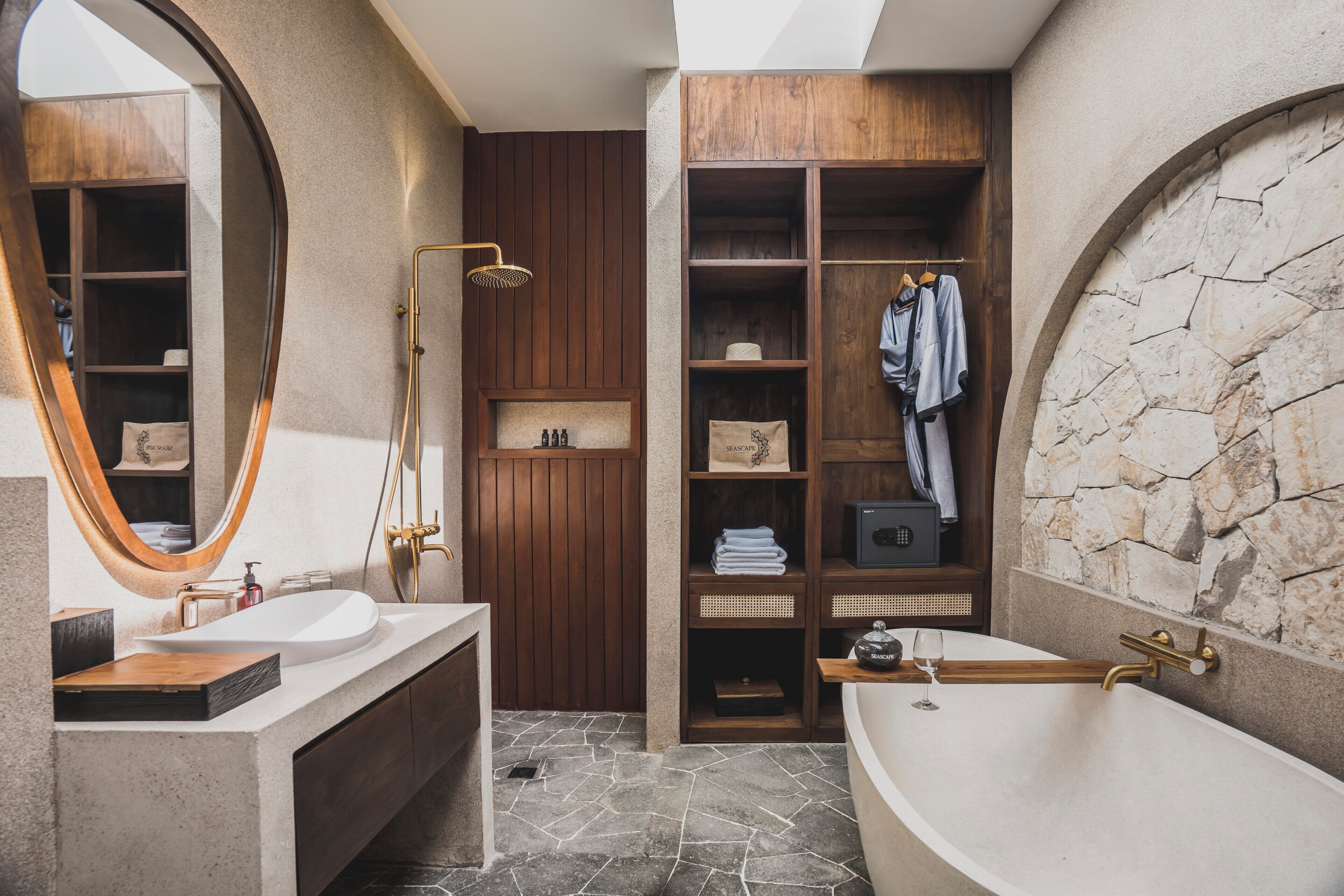 Un baño moderno con una bañera independiente, un espejo grande, estanterías de madera y una pared decorativa de piedra.