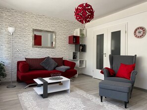 A living room with a red sofa, a grey armchair, a white coffee table, a red pendant light, and a clock on the wall. - Apartment 'Dorfblick' with Private Terrace and Wi-Fi (Olpe)